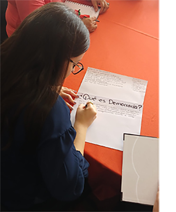 A young woman is writing. The words are "Que es democracia?" or "what is democracy?"