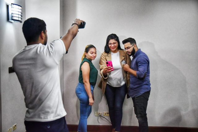 Three young participants stand looking at a mobile phone, while another takes a photo with his phone. 