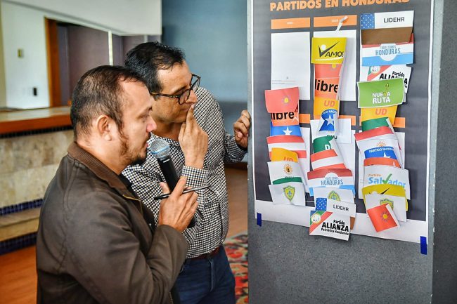 Two participants look at a colourful board filled with the logos or civil society organizations and political parties.